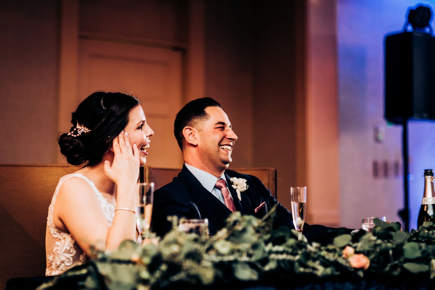 A couple shares a joyful moment at their wedding reception, laughing together at the head table with floral decor.