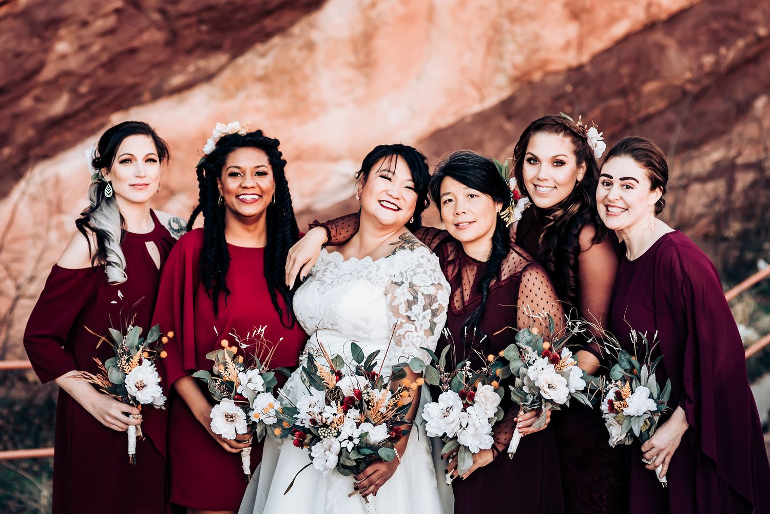 Wedding party with bridesmaids in burgundy dresses holding floral bouquets against red rock backdrop.