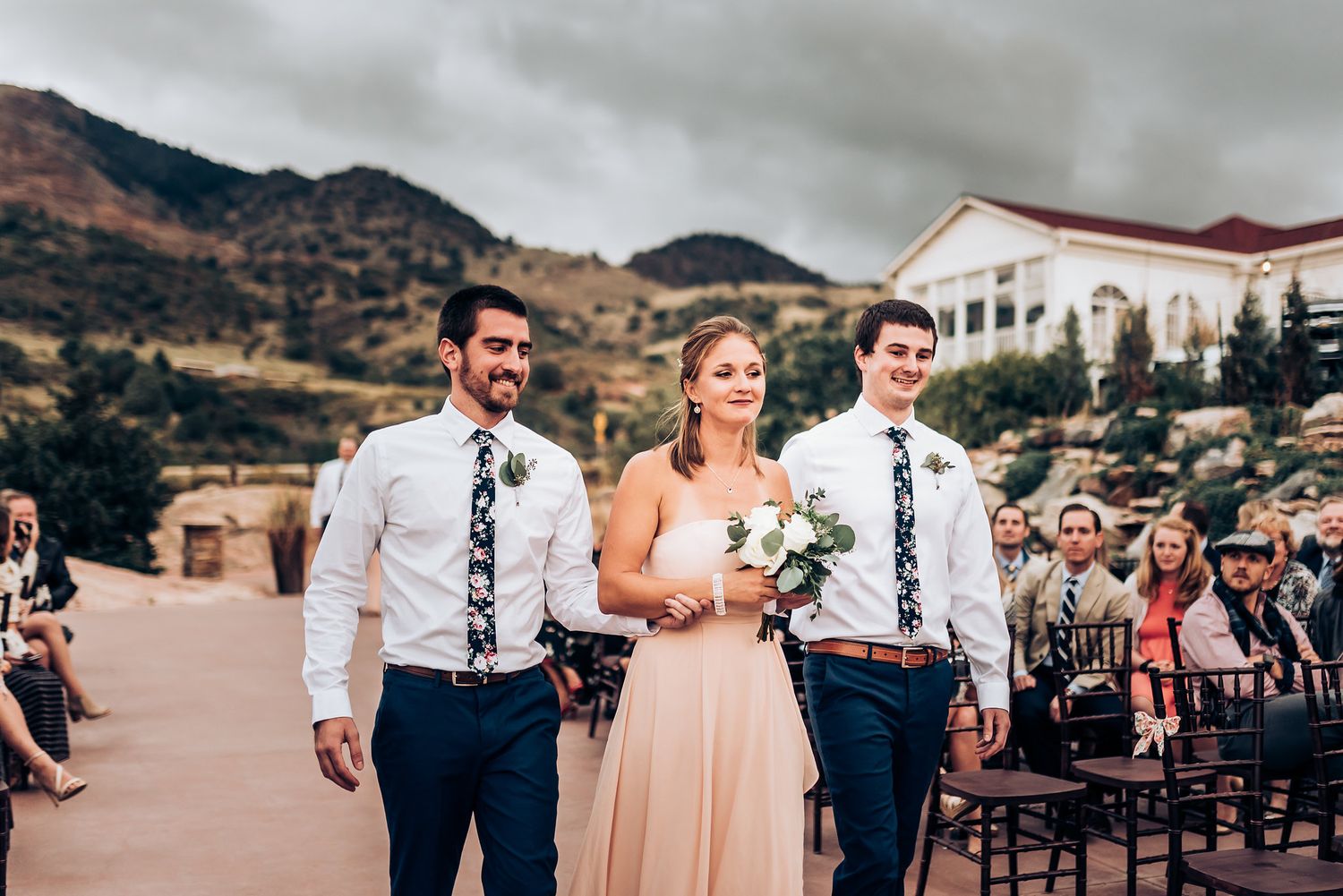 Wedding party members walk down an outdoor aisle with mountain views and guests seated on both sides.