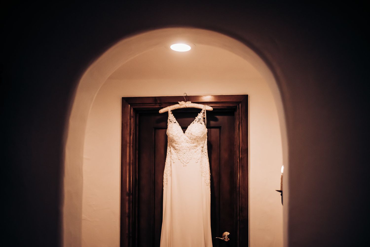 Elegant white wedding dress hanging in an arched doorway with dark wooden door.