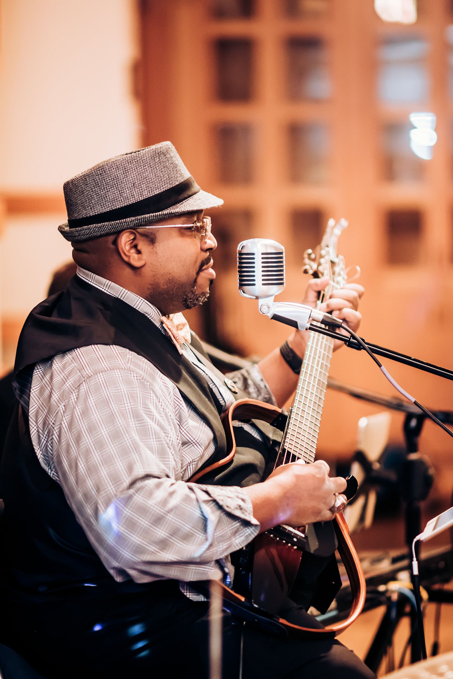 Musician in fedora hat and vest performs with guitar and vintage microphone.
