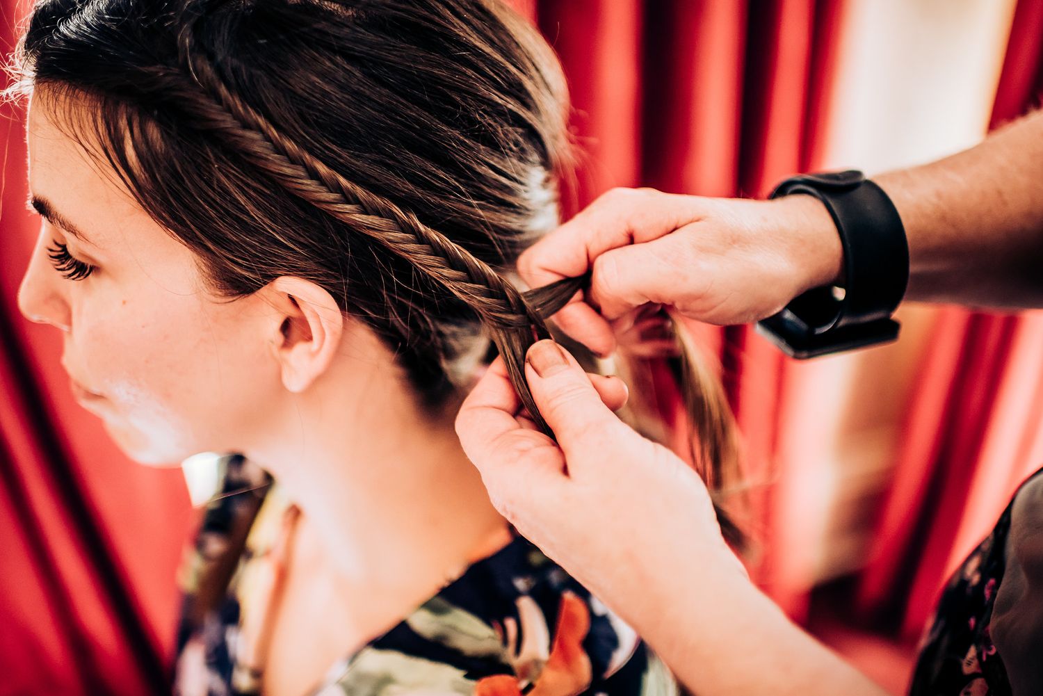 Close up of hands braiding hair against red curtain background.