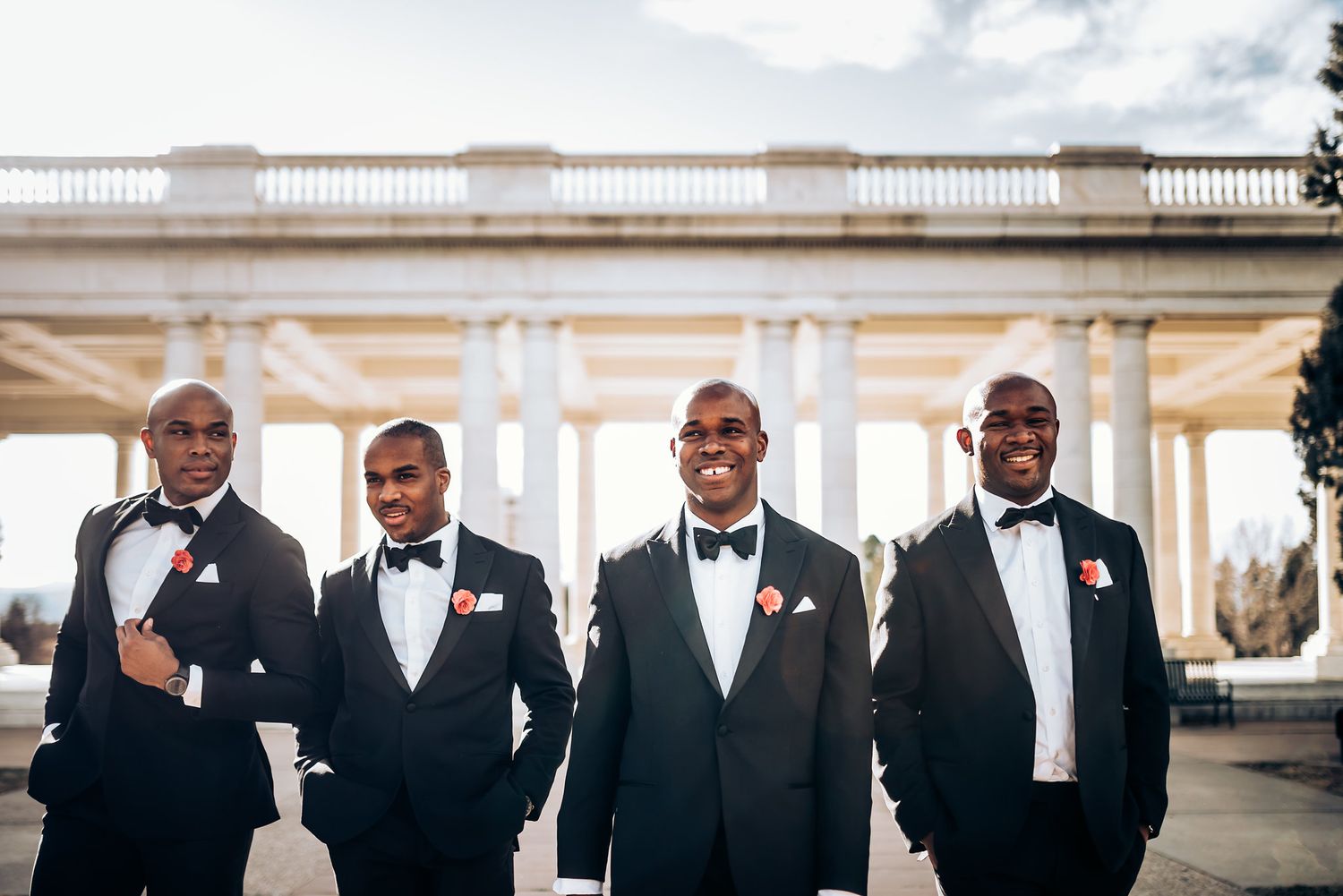 Groomsmen in classic black tuxedos pose confidently against white columned architecture.
