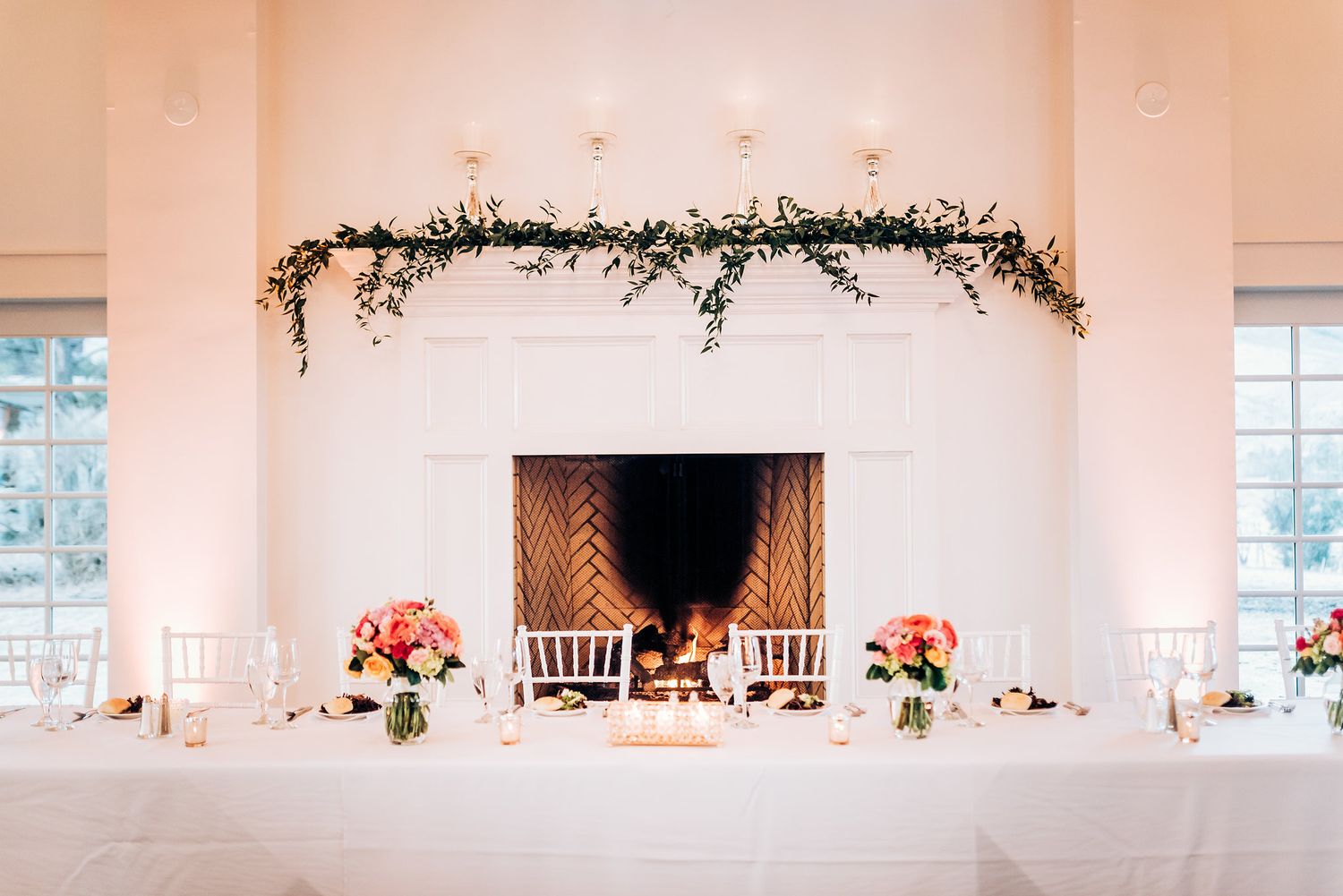 Elegant wedding reception table setup with floral centerpieces and greenery garland above a fireplace.