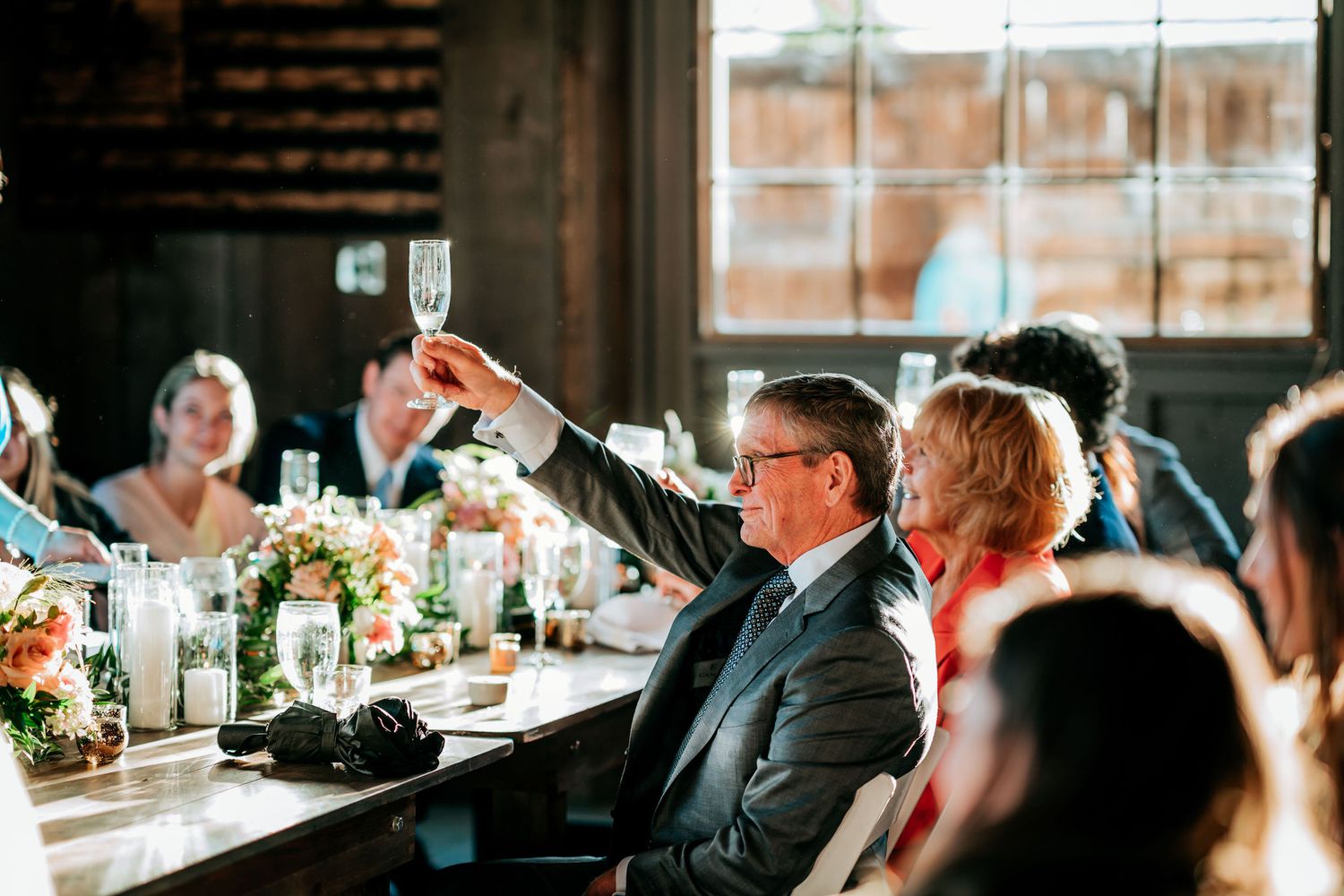 Guests raise their glasses in a celebratory toast during an elegant wedding reception in a rustic venue.