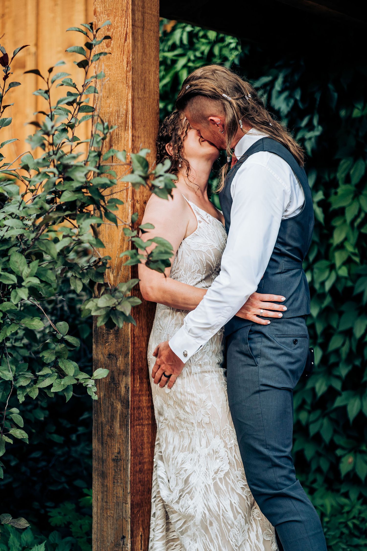 Romantic wedding couple embraces near wooden post surrounded by green foliage.