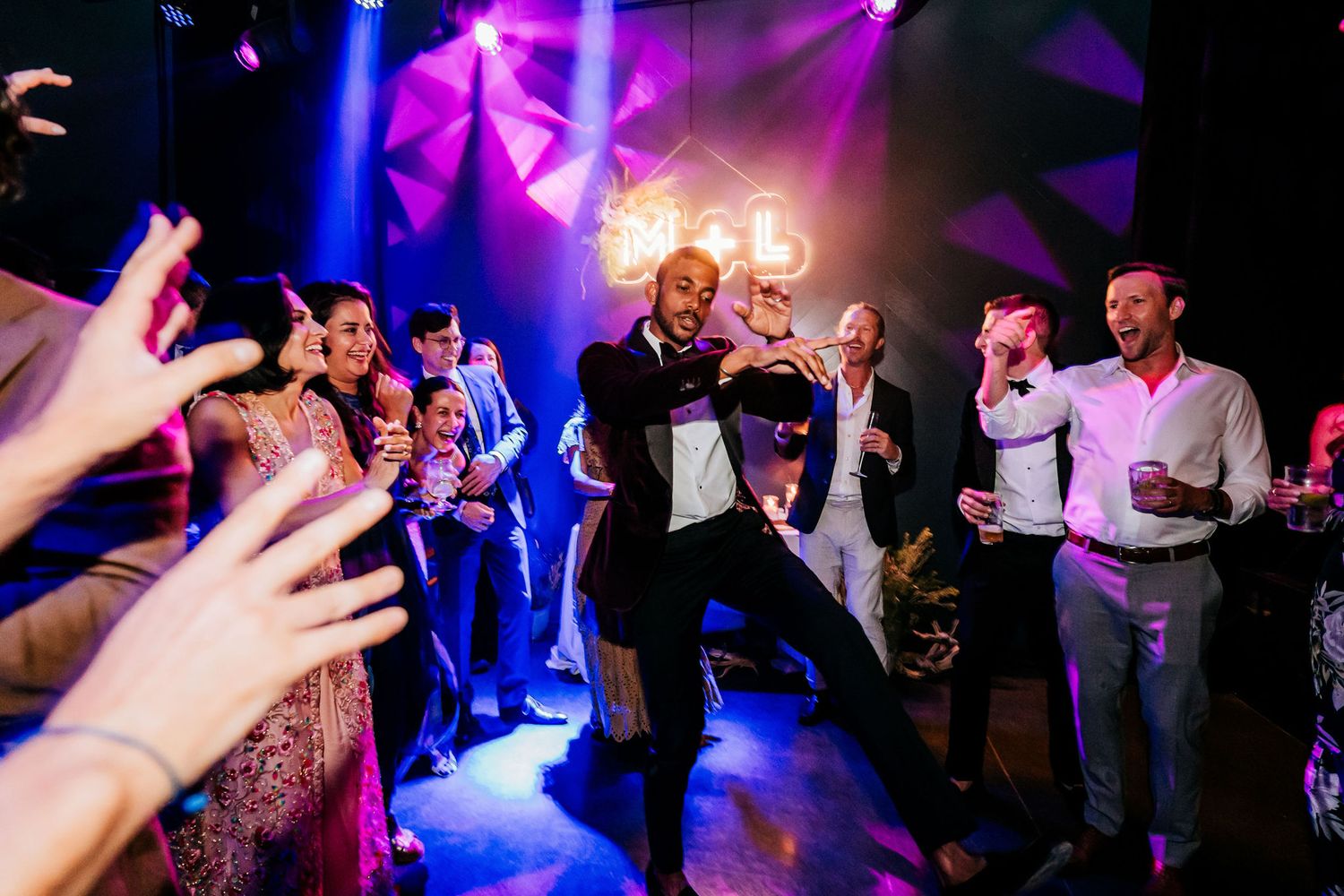 Wedding guests dancing enthusiastically under vibrant purple lighting at a reception party.