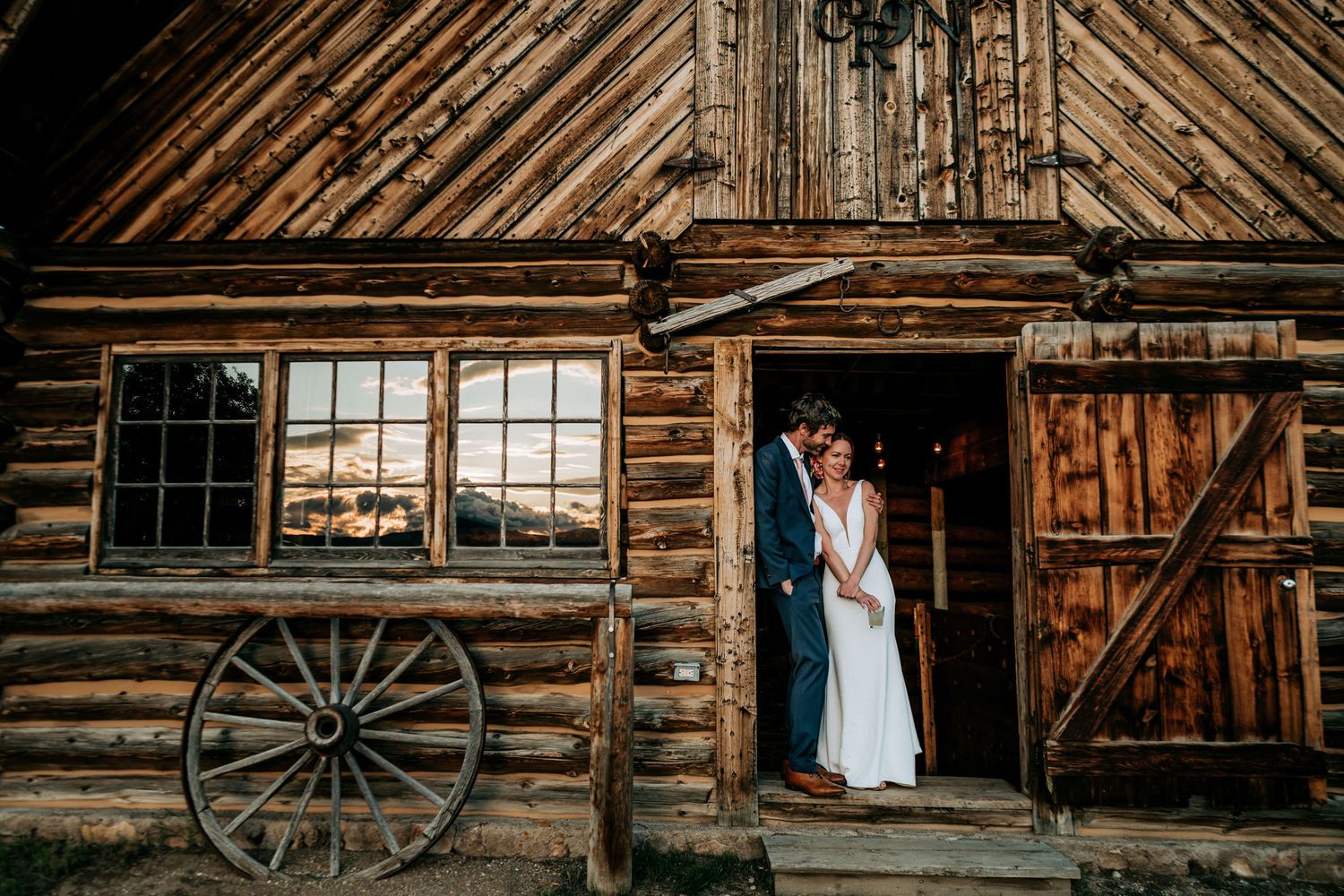 A couple poses in the doorway of a rustic wooden barn with an antique wagon wheel.