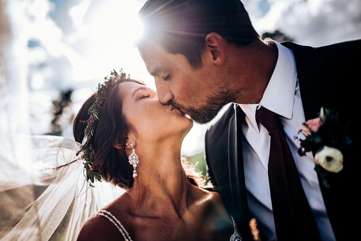 Backlit wedding portrait captures intimate kiss with flowing veil and formal attire.