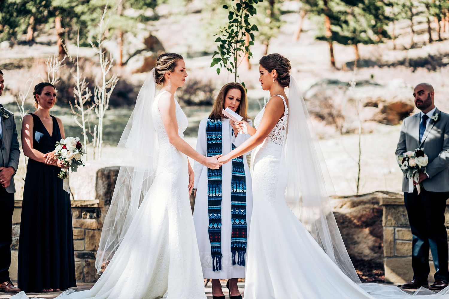 A wedding ceremony takes place outdoors with two brides in white dresses and an officiant in blue standing under a wooden arch.