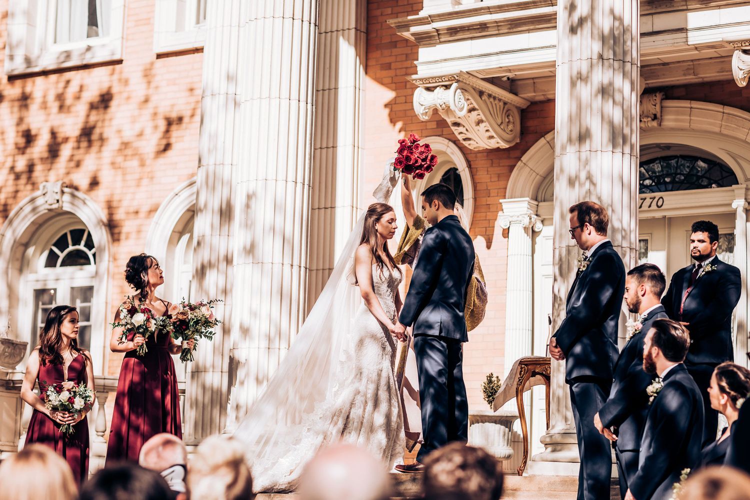 Wedding ceremony taking place on elegant stone steps of historic building with white columns in warm sunlight.
