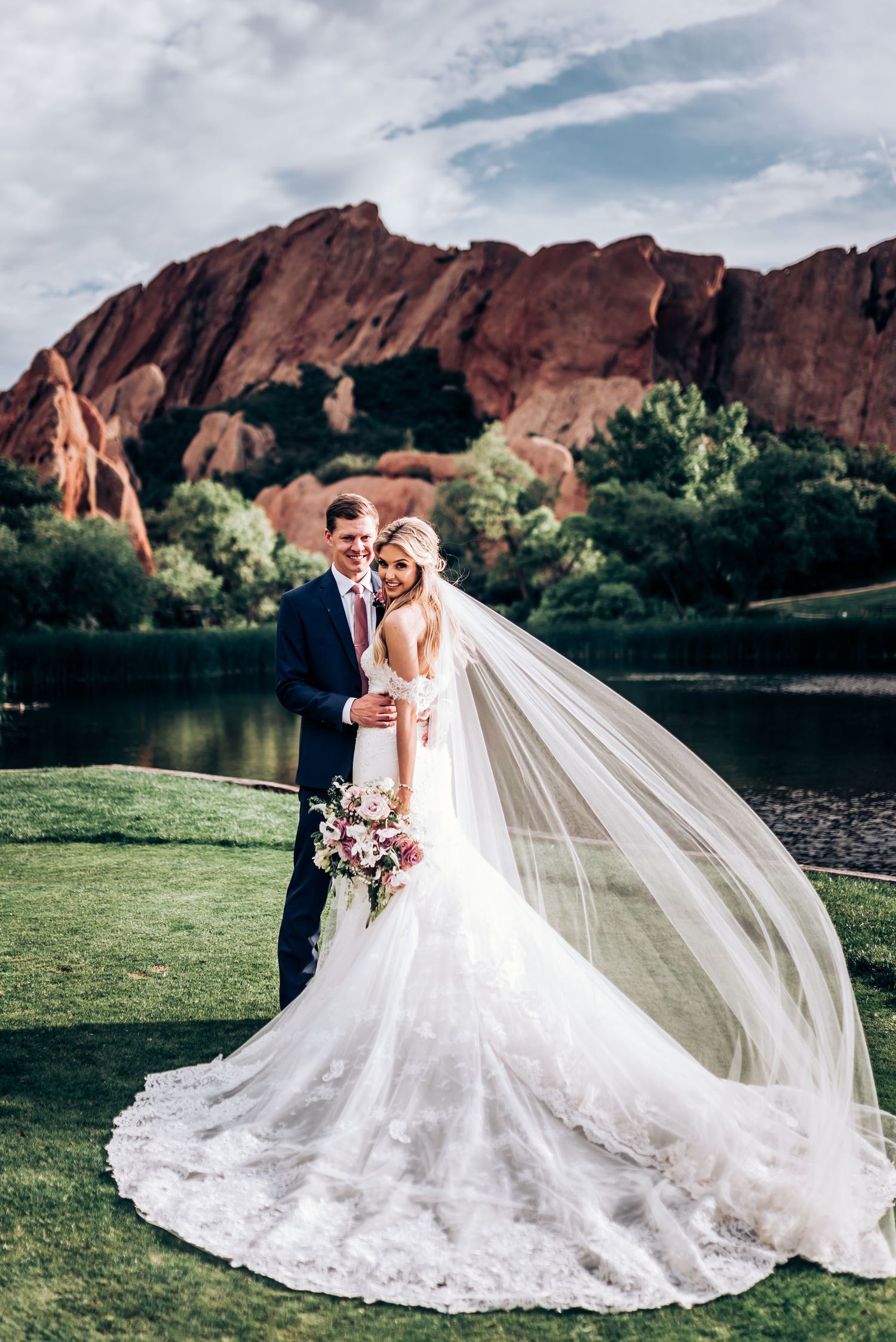 Elegant wedding photo showcasing flowing veil against remarkable red rock formations and serene lake.