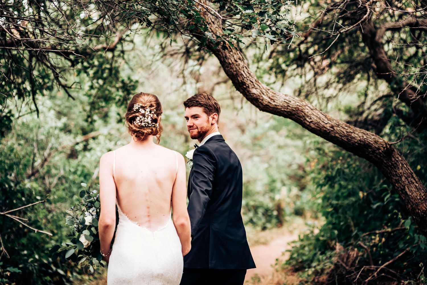 Elegant couple in wedding attire pose beneath a twisted tree branch on a nature path.