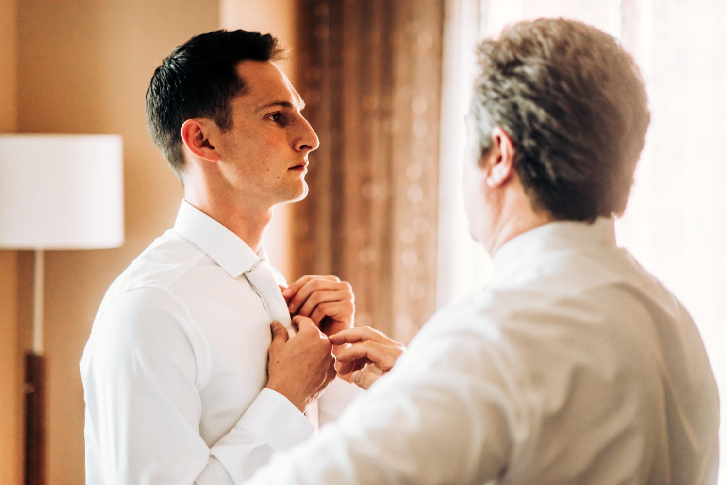 Two people in white dress shirts adjusting formal attire in a warmly lit room.