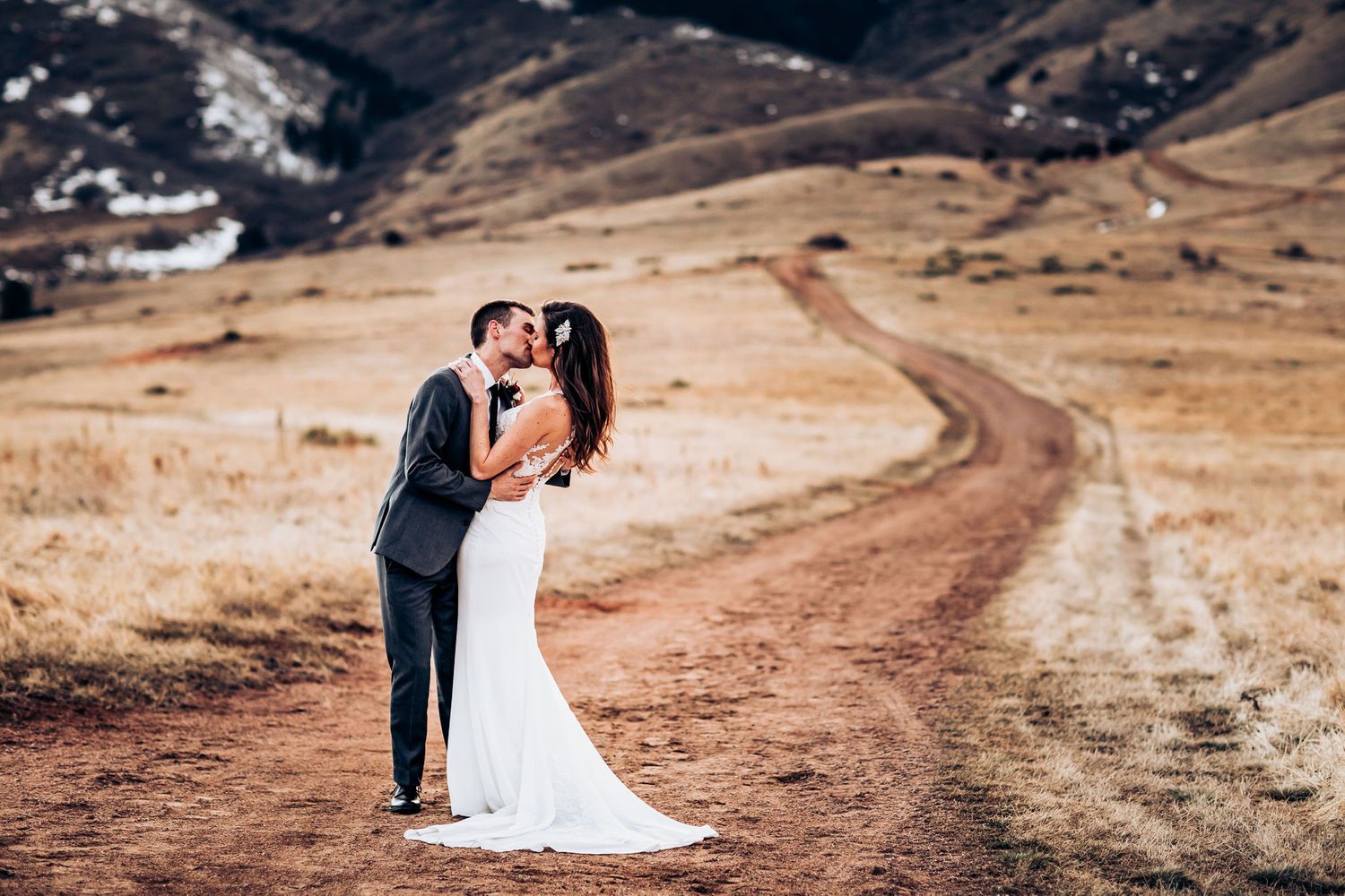 Romantic wedding portrait of couple embracing on dirt path with mountains in background.