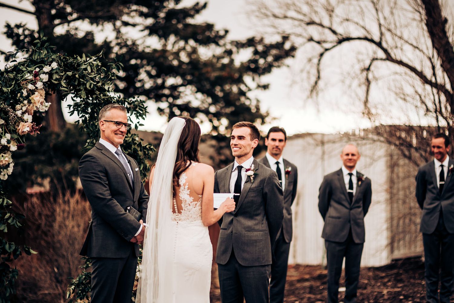 Wedding party stands beneath evergreen branches during outdoor ceremony.