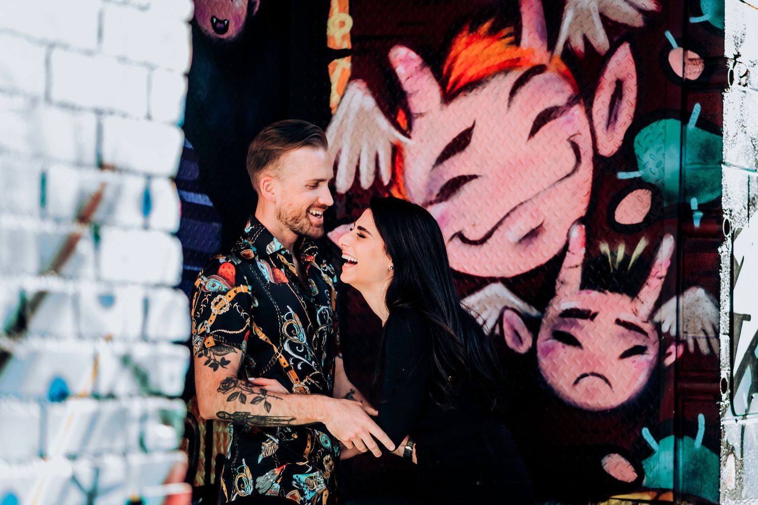 A couple laughing together against a wall decorated with pink street art.