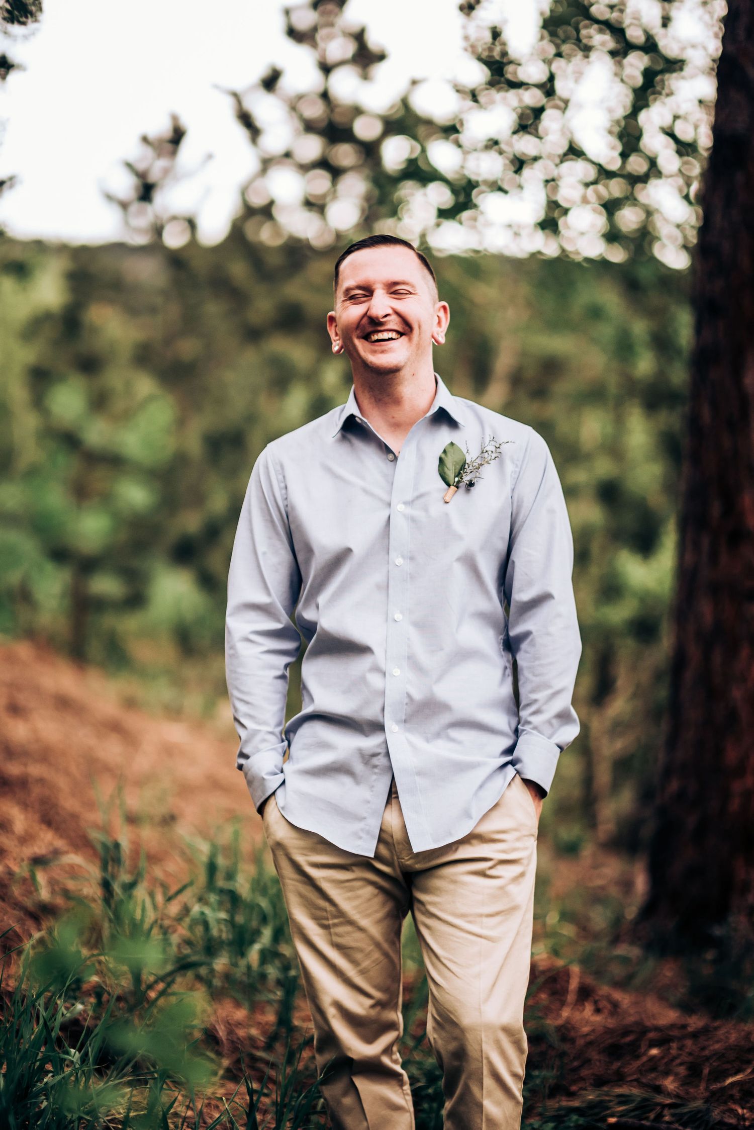 Groom in light grey shirt and khakis smiles in outdoor forest setting.