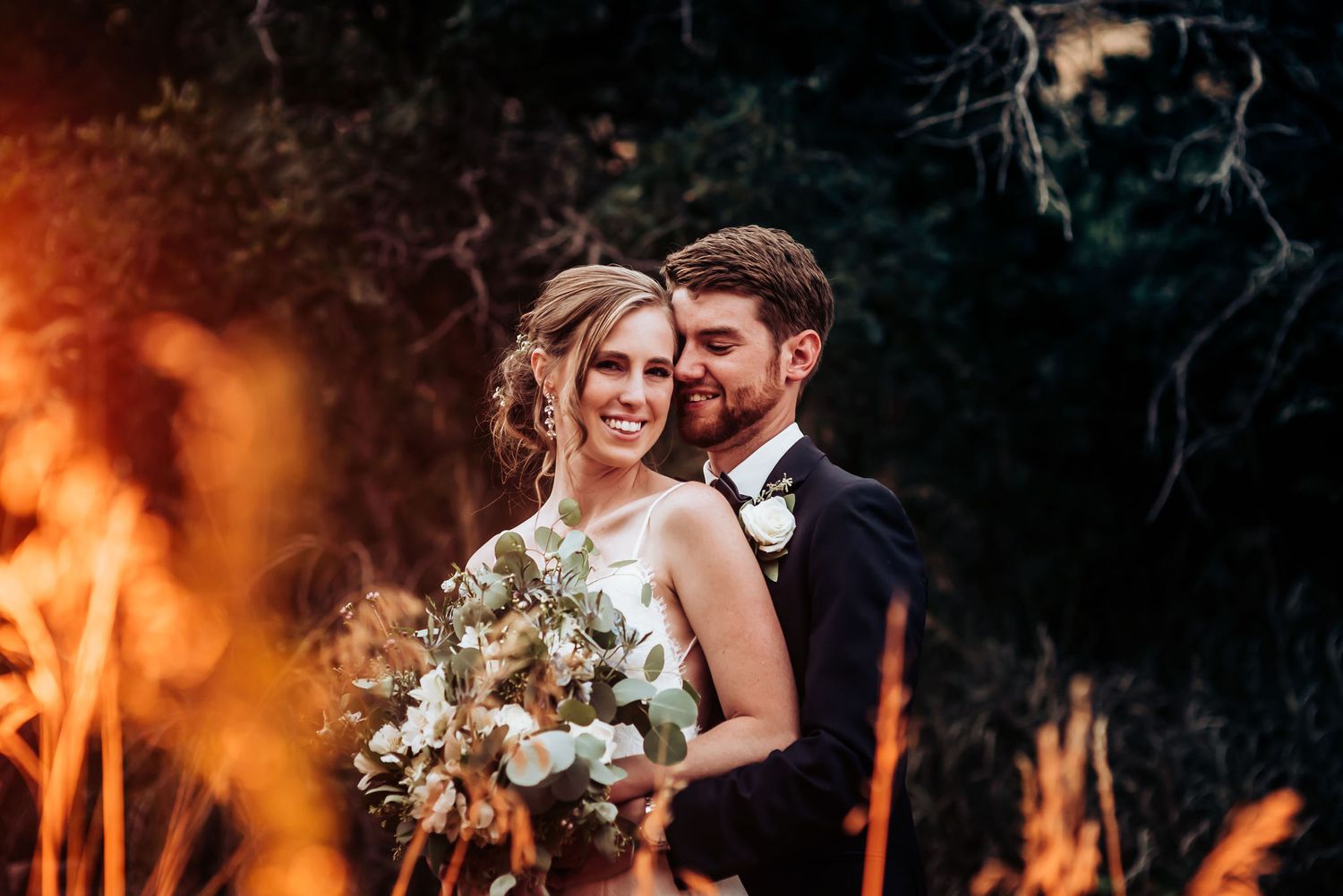 Dramatic wedding portrait with glowing sunset and bouquet of white flowers.