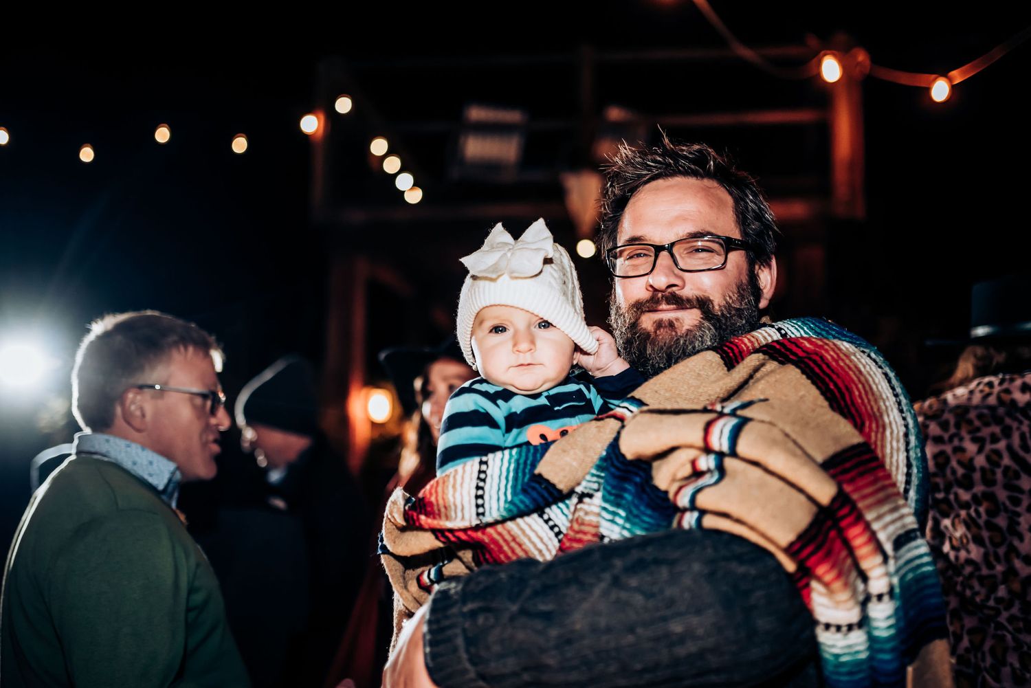 A baby in winter attire is held while wrapped in a striped blanket at a nighttime gathering.