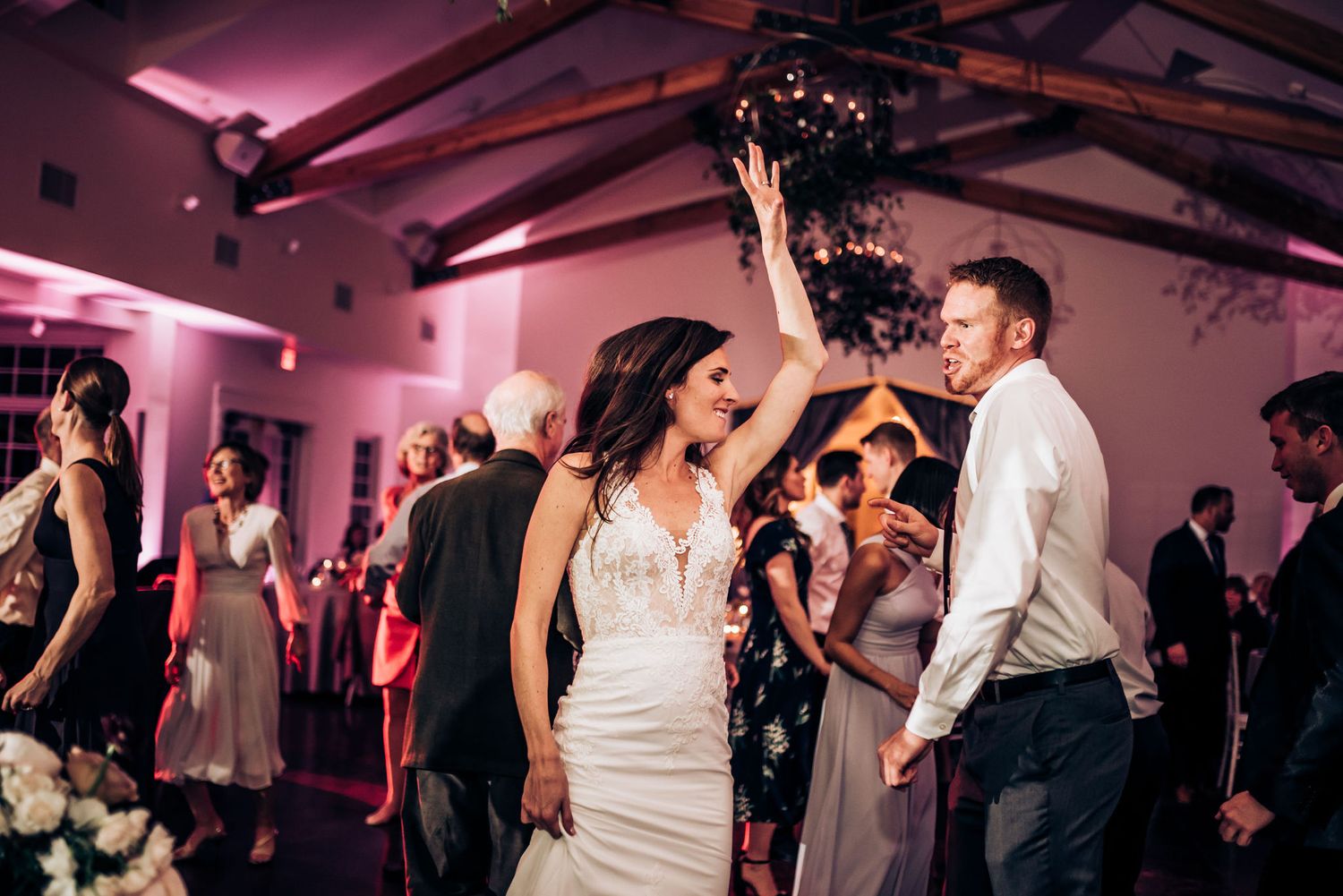 Wedding celebration in full swing with guests dancing under exposed wooden beams and pink lighting.