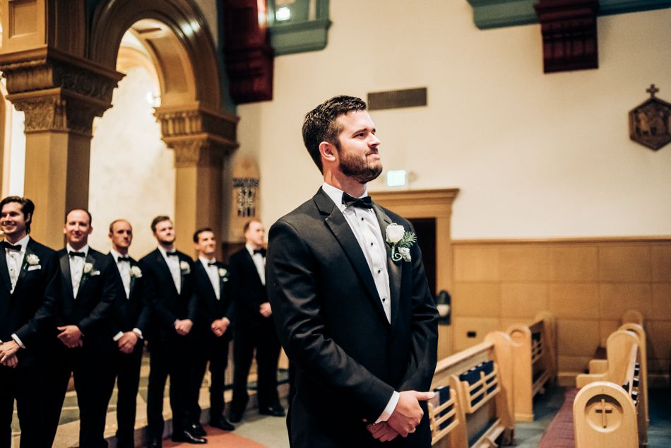 Groom and groomsmen in classic black tuxedos stand in traditional church setting.