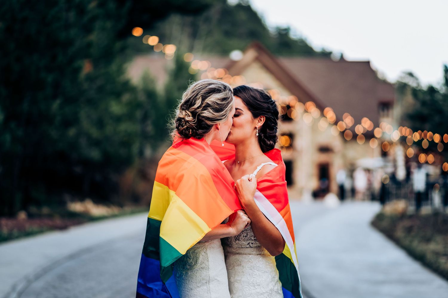 Two people wrapped in rainbow flags share a joyful moment in their wedding dresses.