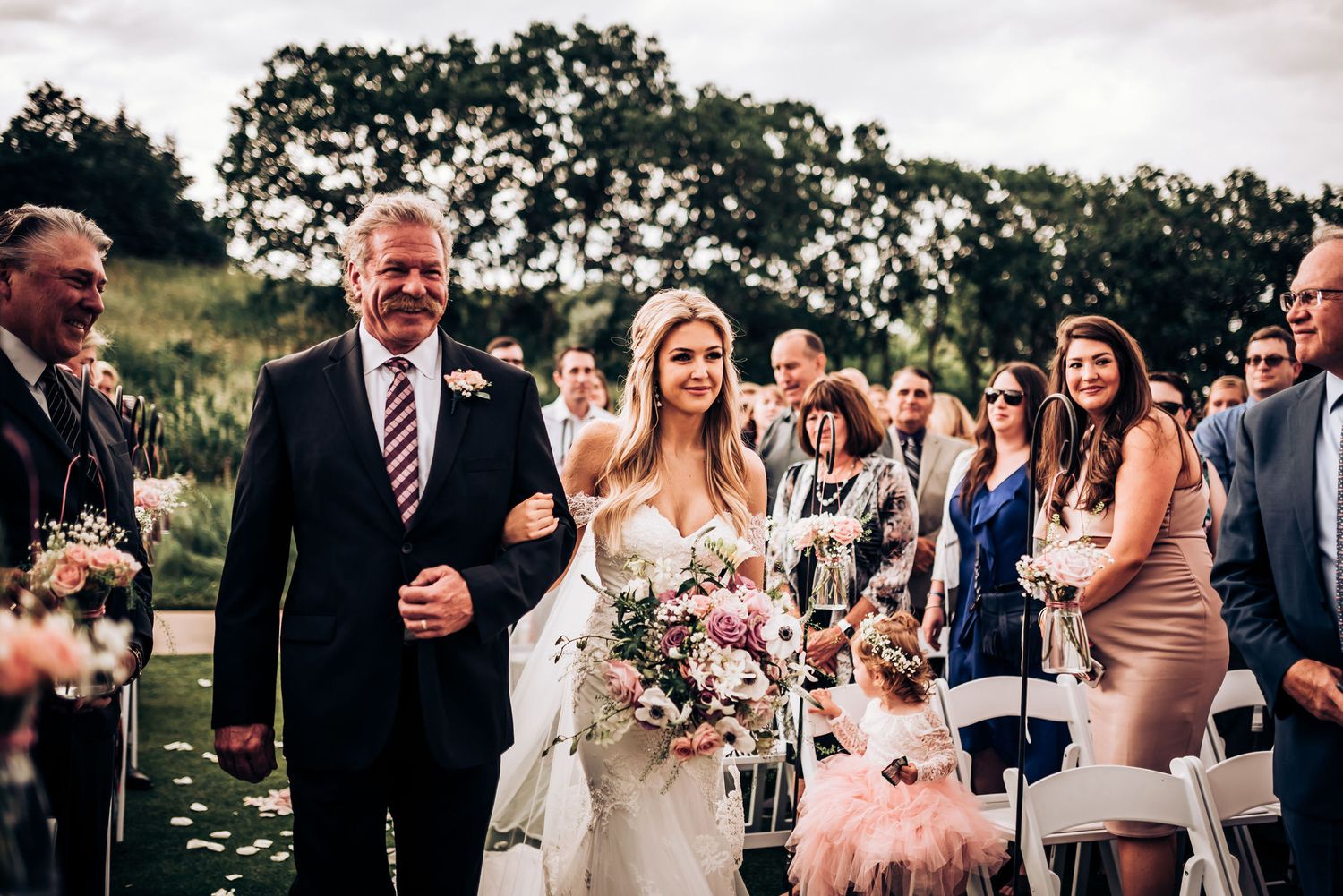 Wedding guests smile and celebrate during an outdoor ceremony as a person carries a beautiful floral bouquet.