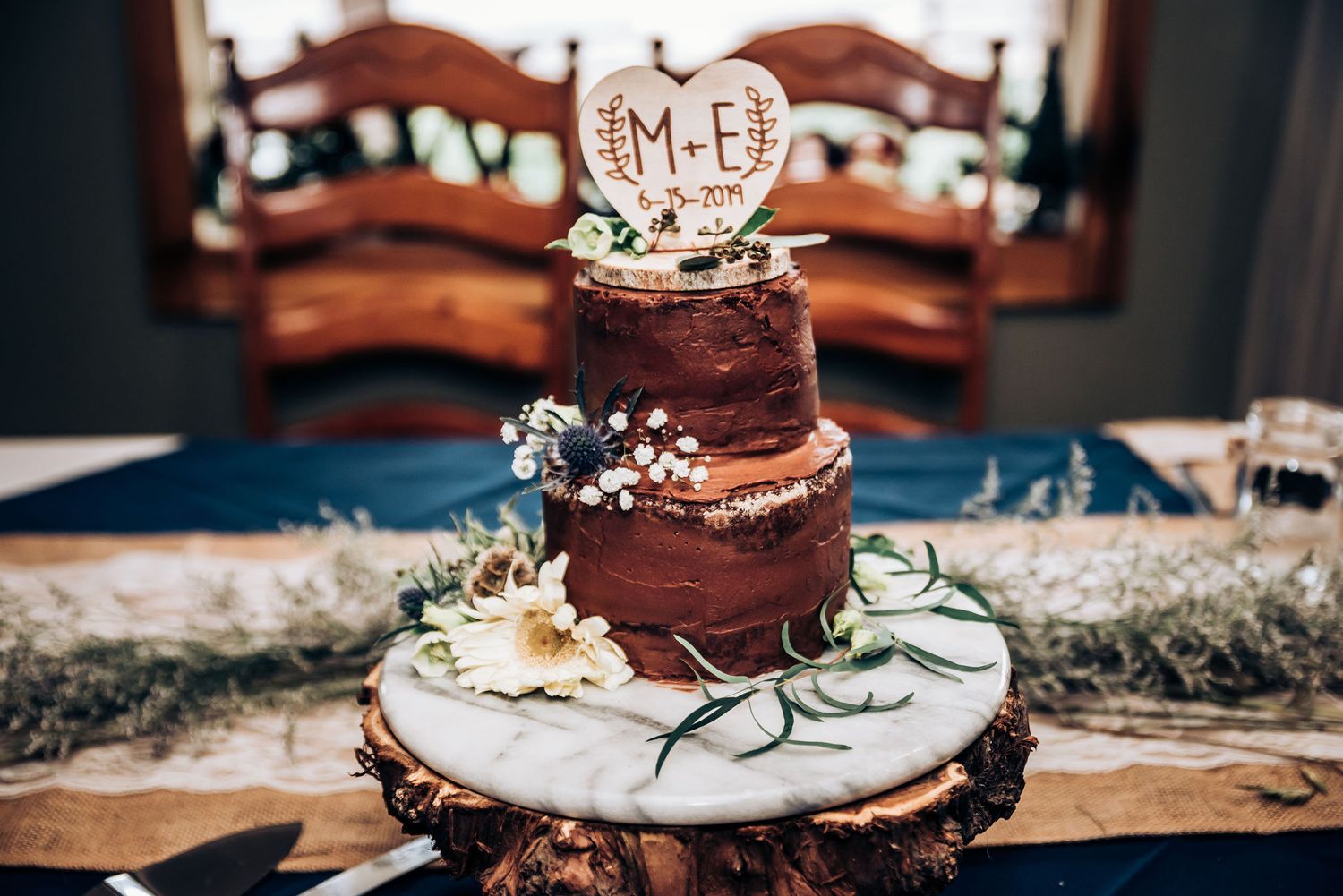 Three-tiered chocolate wedding cake decorated with white flowers and personalized topper on rustic stand.