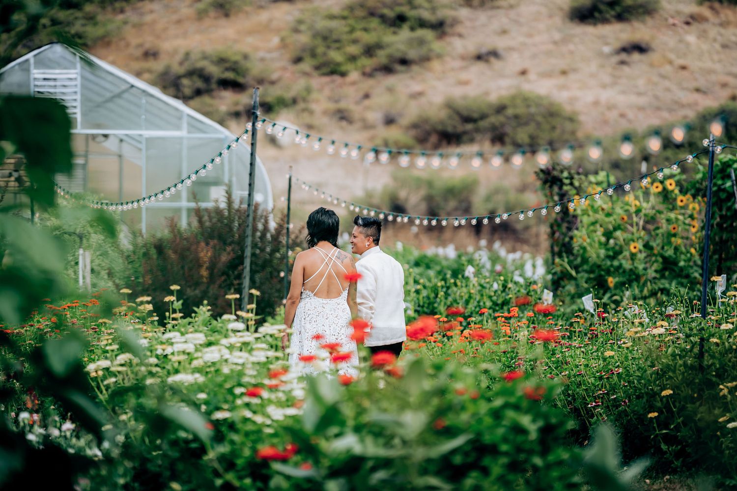 Two people sit among colorful flowers in a garden with string lights overhead.