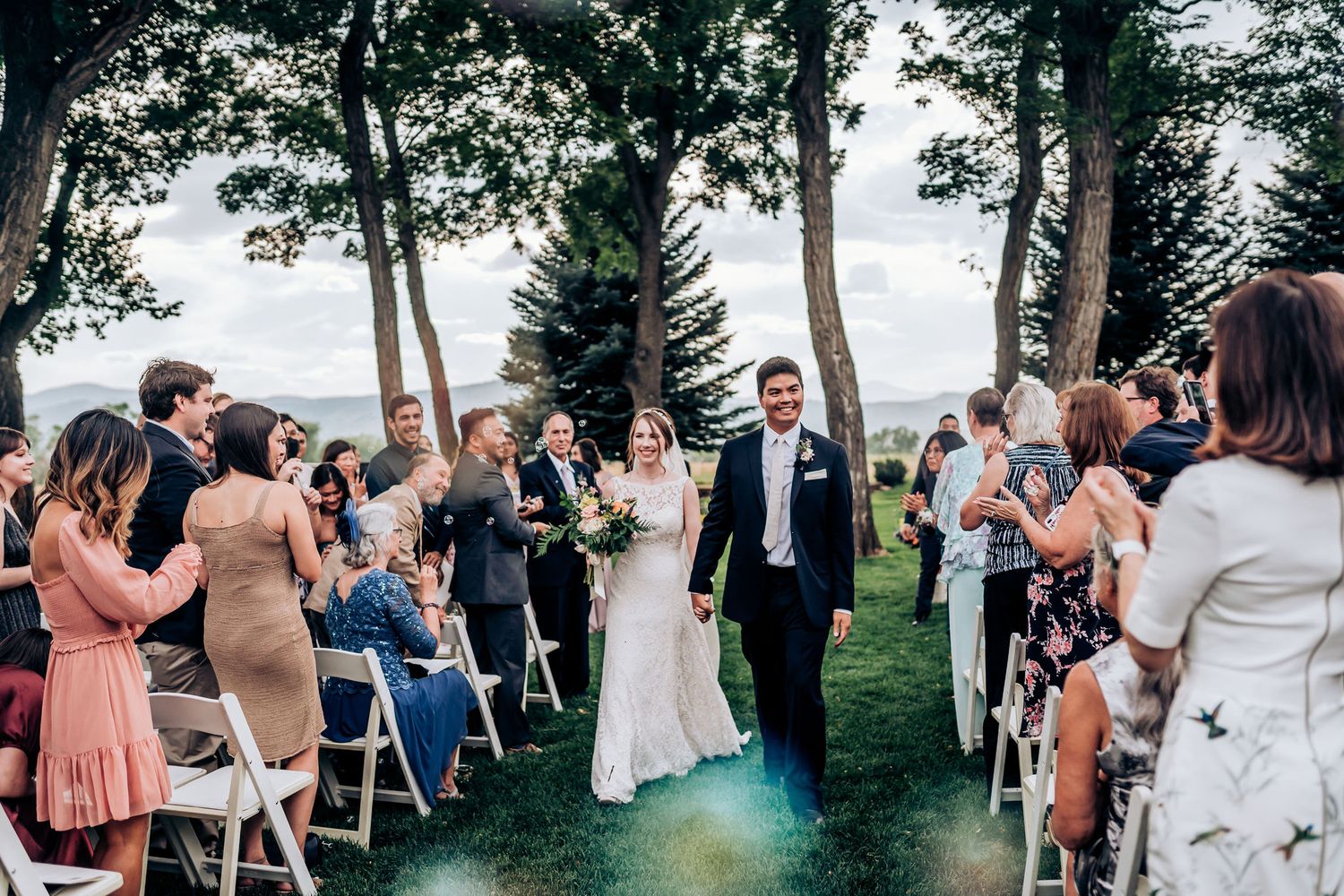 Newlyweds walk down the aisle while guests stand and celebrate at an outdoor forest wedding.
