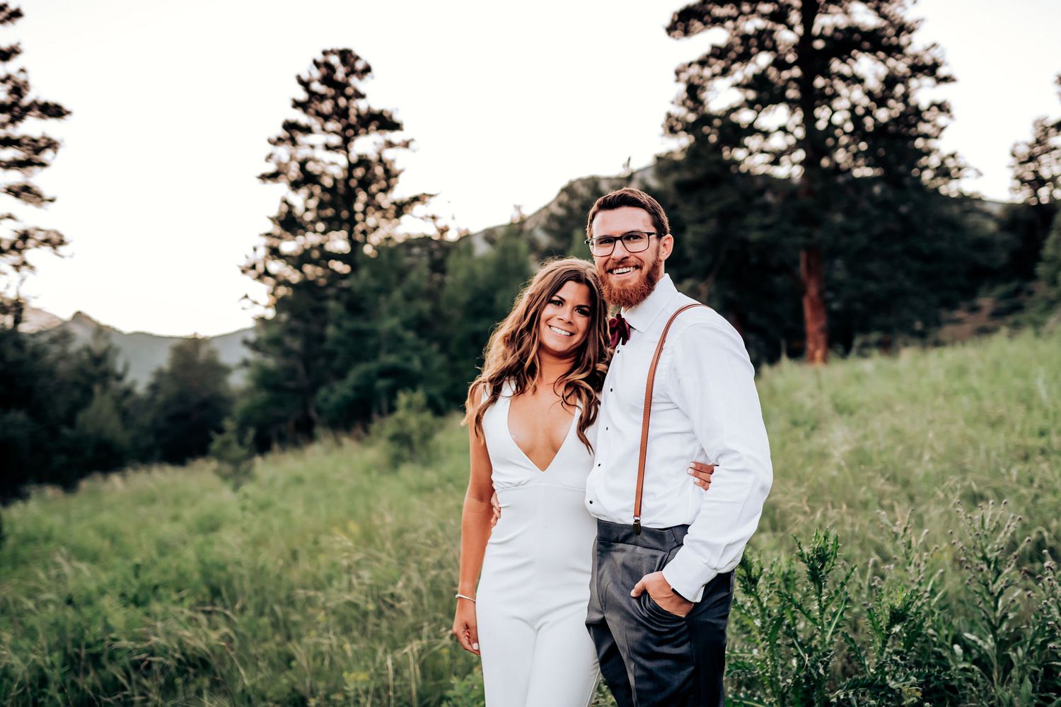 A couple stands together on a grassy hillside with tall pine trees in the background at sunset.