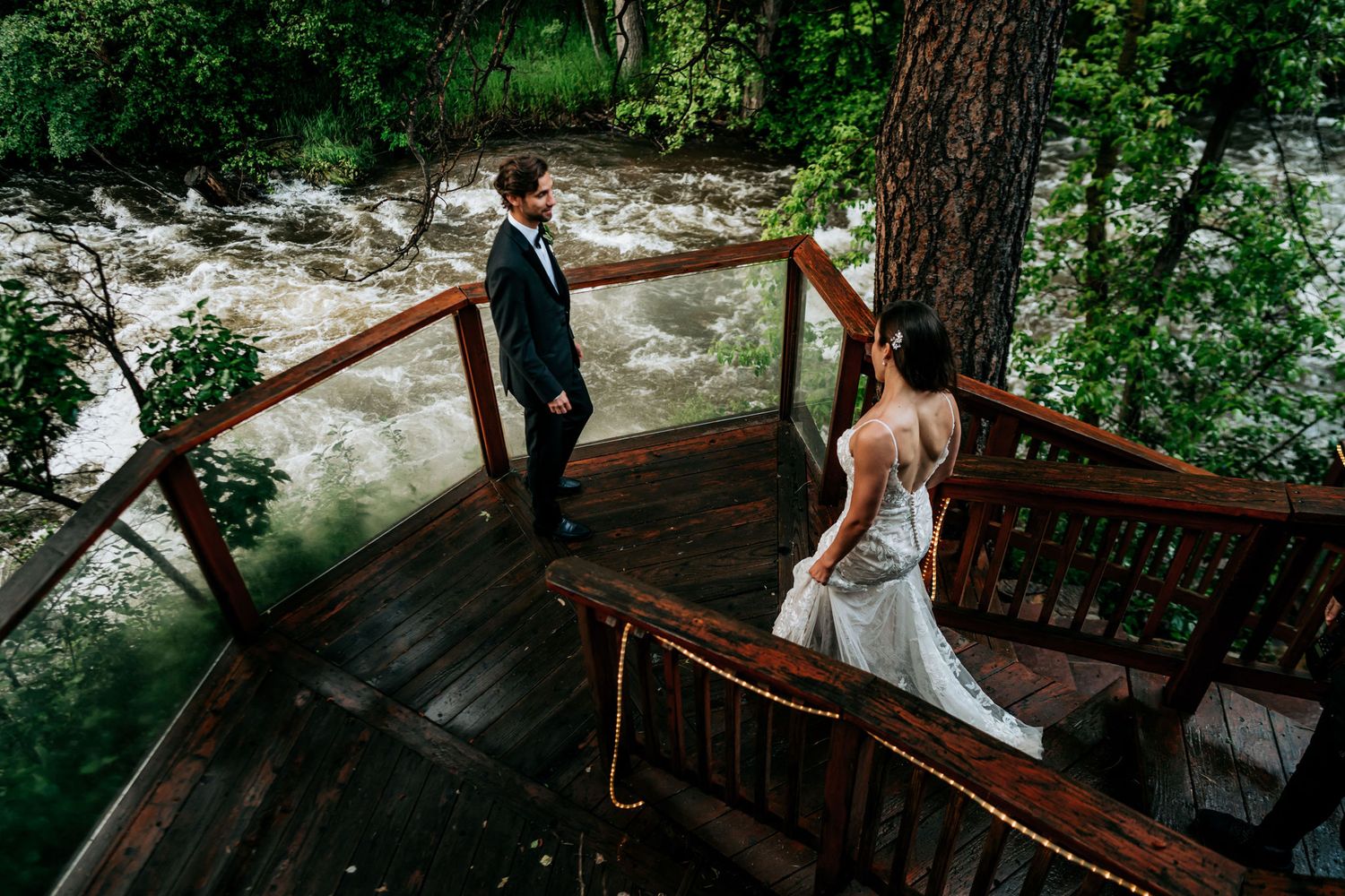 A newlywed couple poses on a wooden deck overlooking a rushing river amid lush greenery.