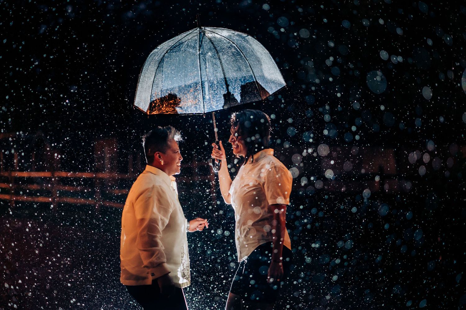 Two people share a romantic moment under a clear umbrella in the rain at night.