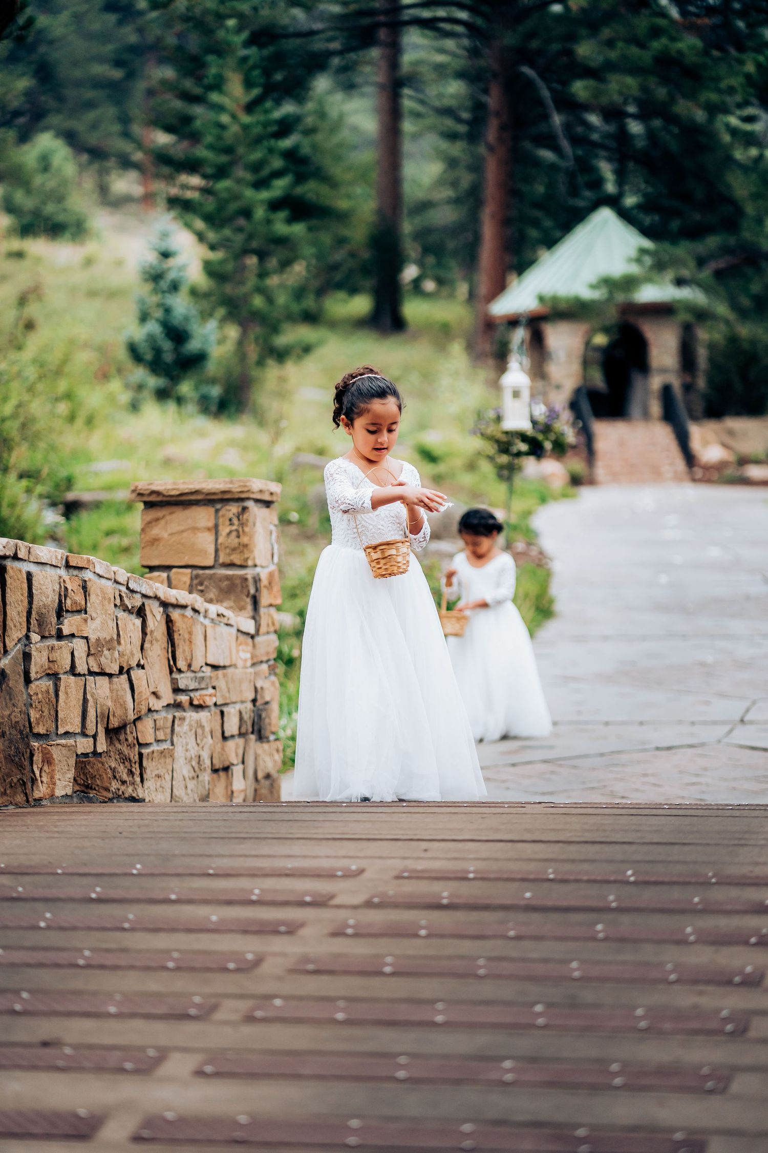 Young attendants in white dresses scatter petals on a wooden walkway with mountain scenery.