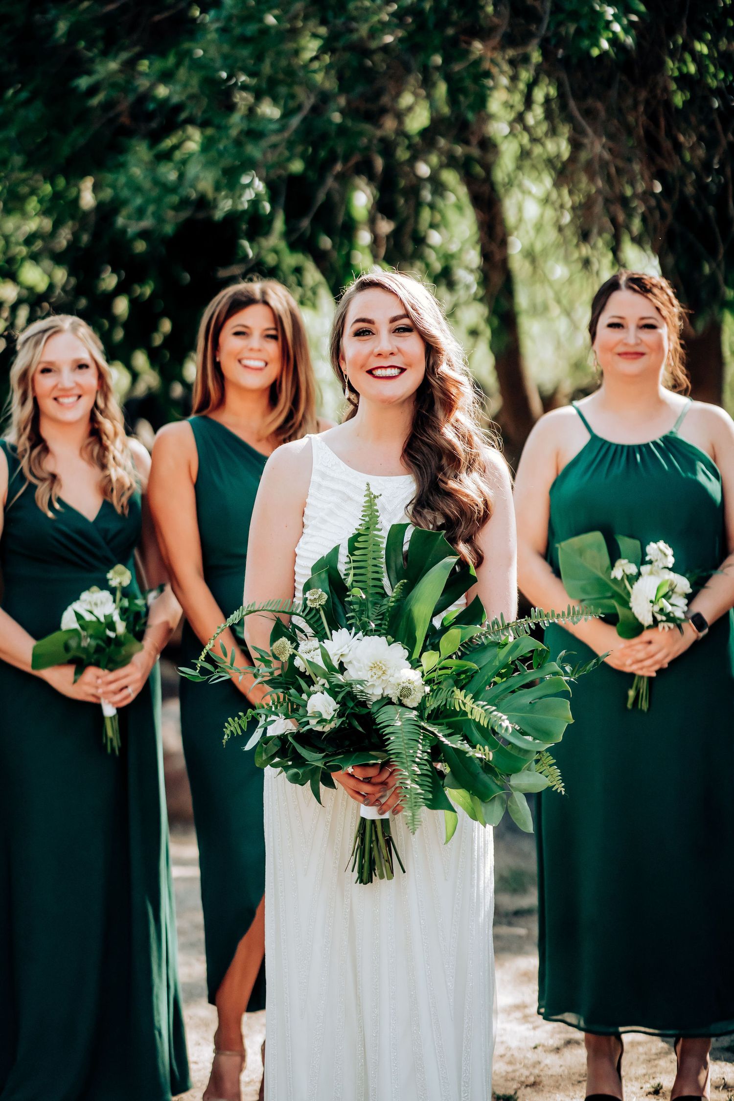 Wedding group in emerald and white poses with tropical leaf bouquets in a sunlit garden.