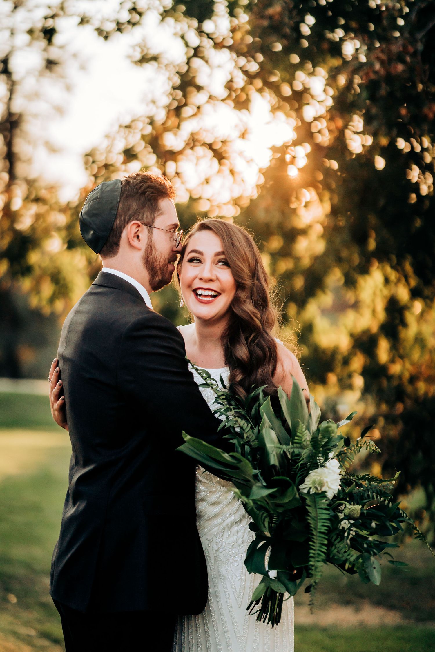 A joyful wedding couple embraces outdoors during golden hour with a lush green botanical bouquet.