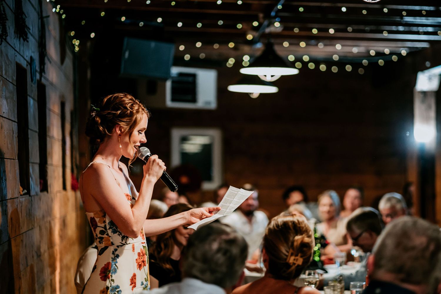 Someone in a floral dress gives a speech at an indoor reception with string lights overhead.