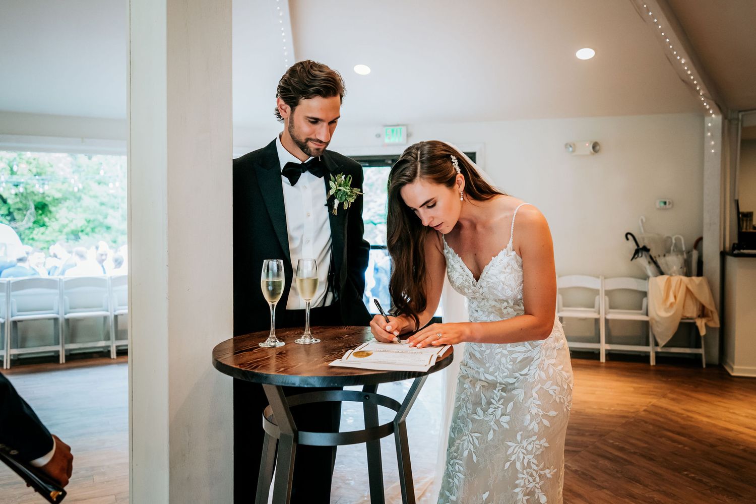 A couple signs documents on a wooden table during a wedding reception.