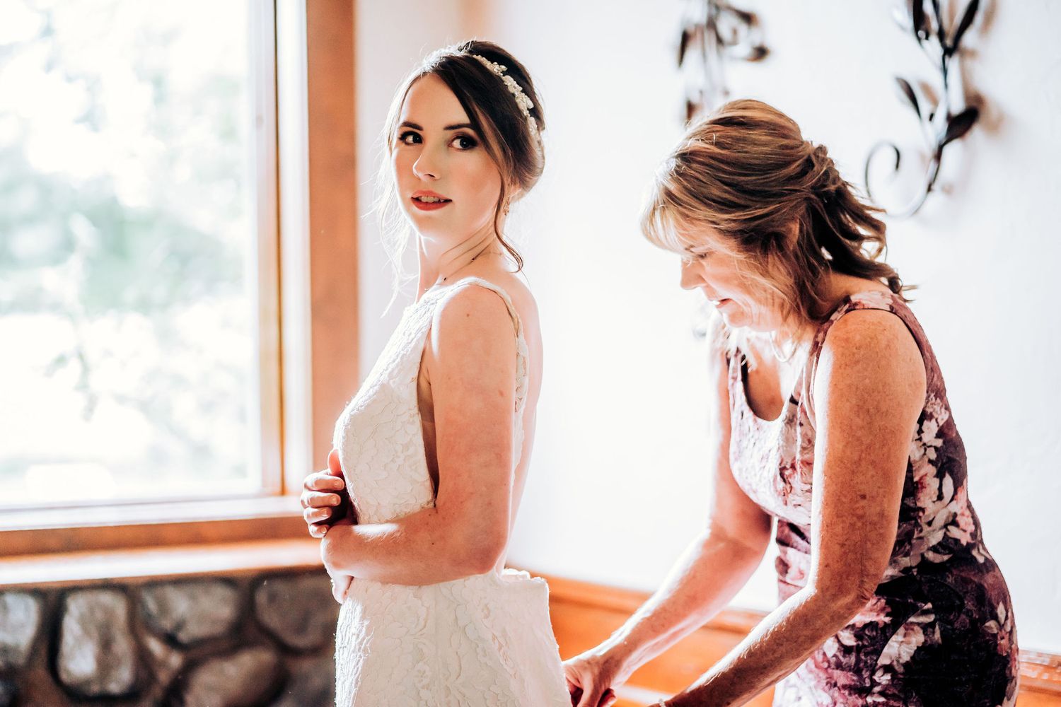 Two people in white dresses prepare for wedding ceremony near window.