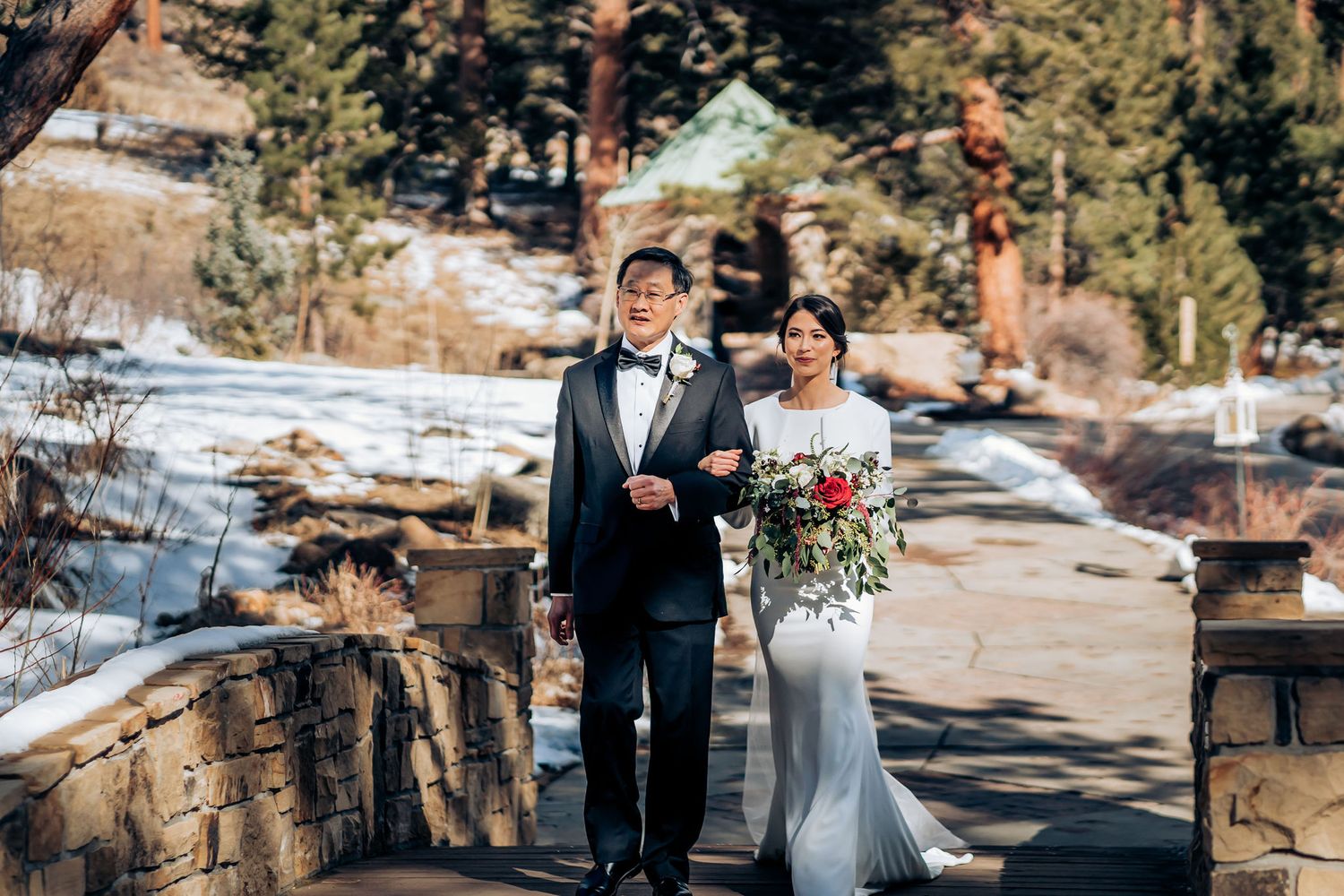 Wedding processional with couple walking along snowy stone path surrounded by pine trees.