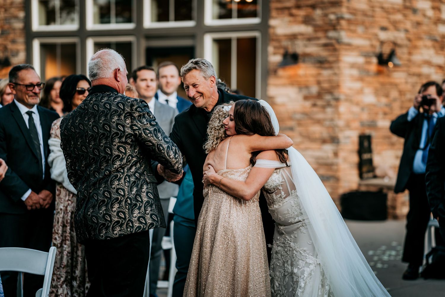 Bride in white veil hugs mother in gold dress at outdoor wedding ceremony.