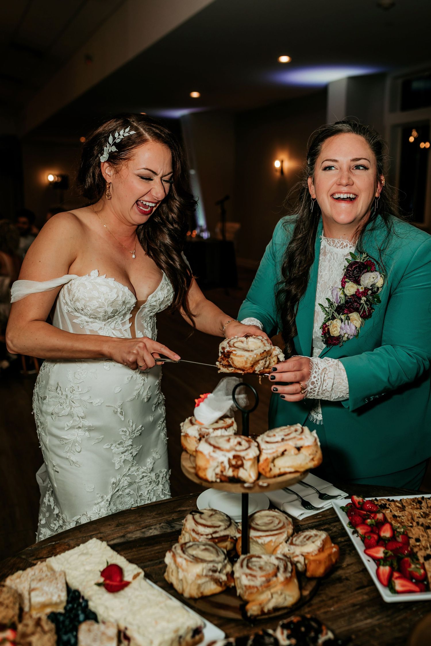 Two brides laughing while cutting cinnamon roll wedding cake at reception.