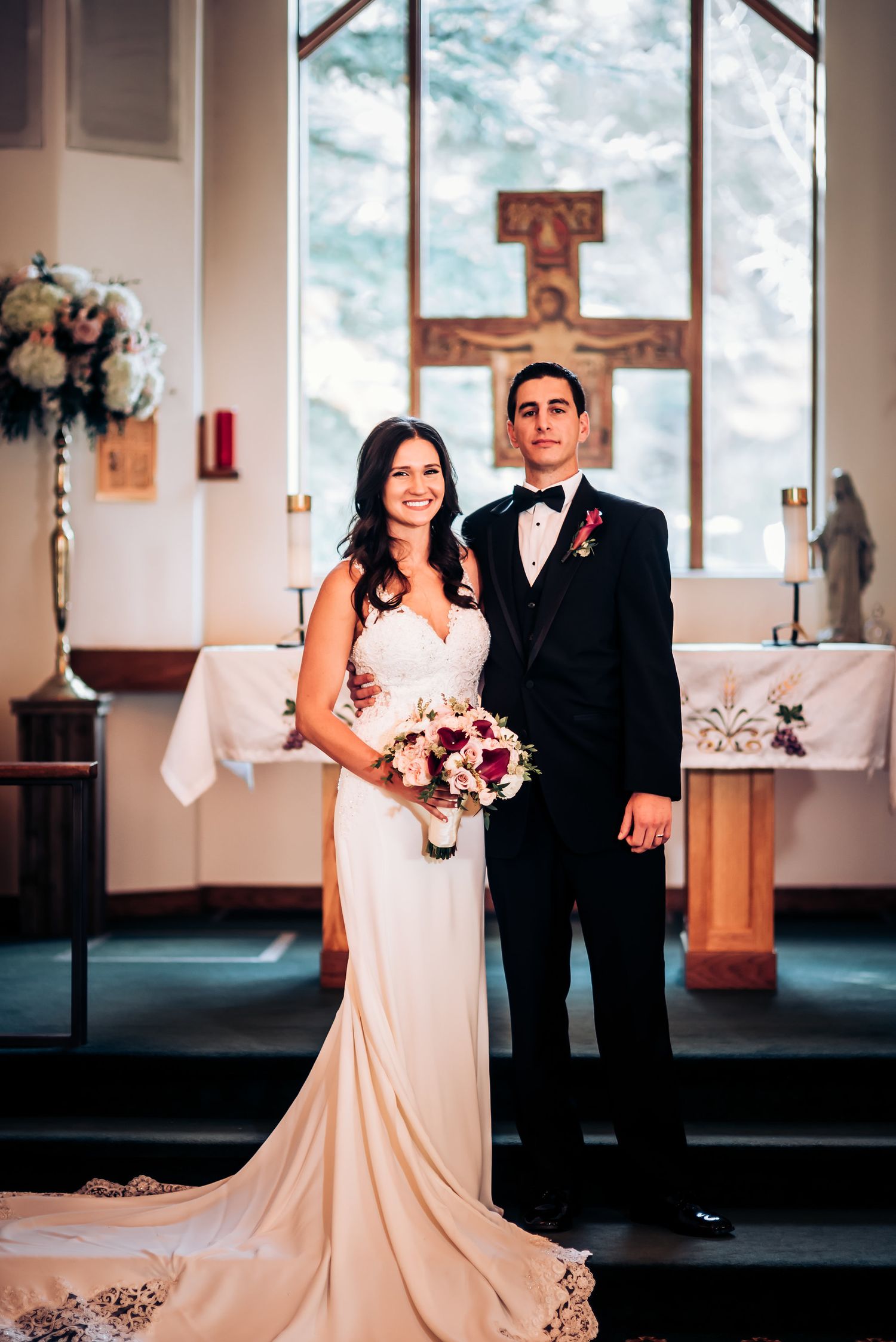 Wedding couple poses in front of church altar with large wooden cross and floral arrangements.