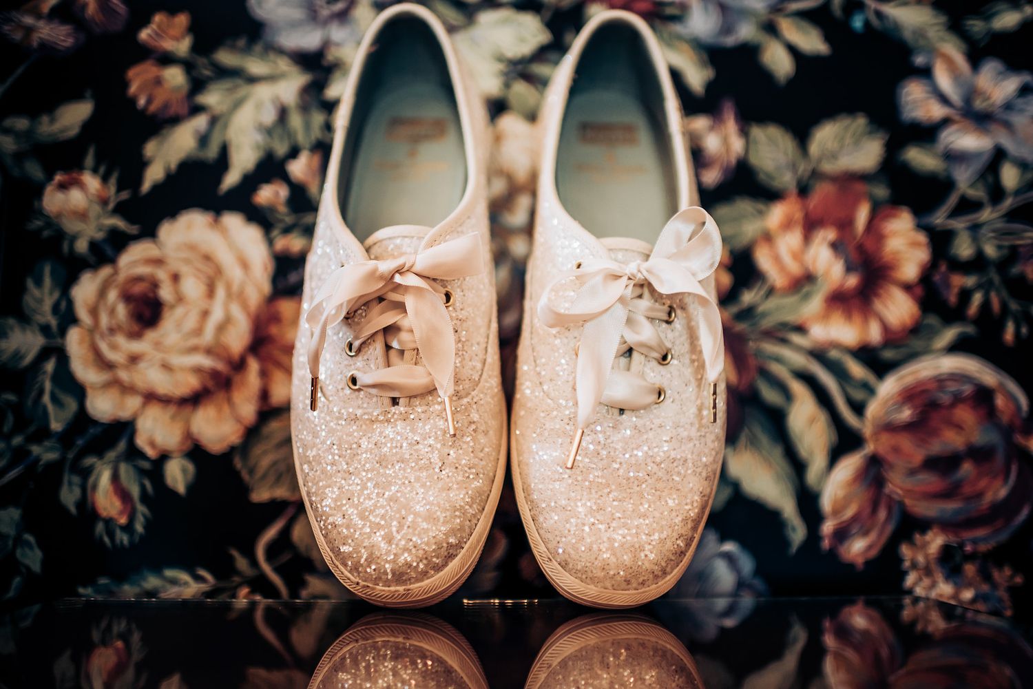 Sparkly white lace-up sneakers positioned on a floral patterned surface.