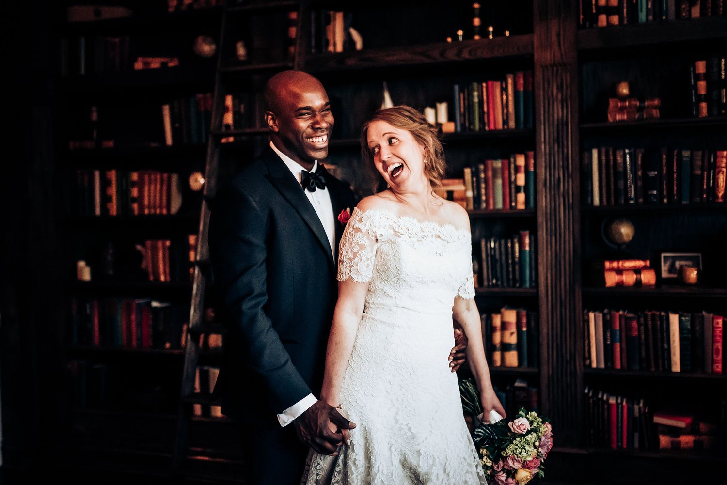Wedding couple posing in a romantic library setting with floor-to-ceiling bookshelves.