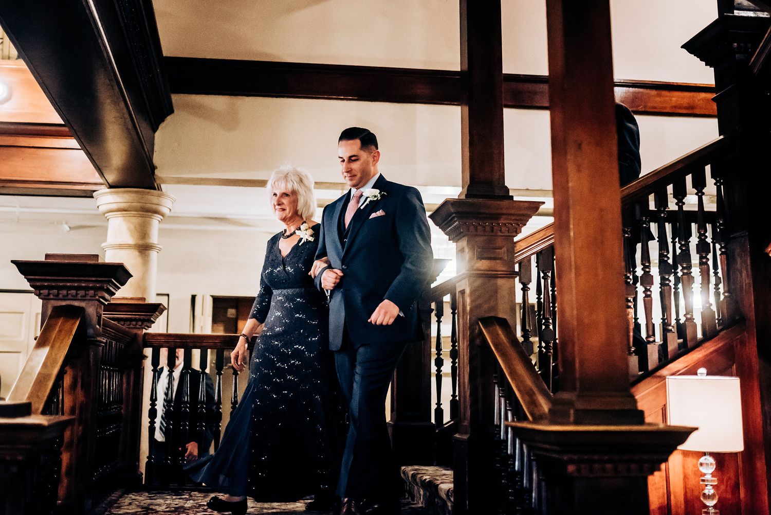 A well-dressed couple stands elegantly on a grand wooden staircase in an ornate historic building interior.