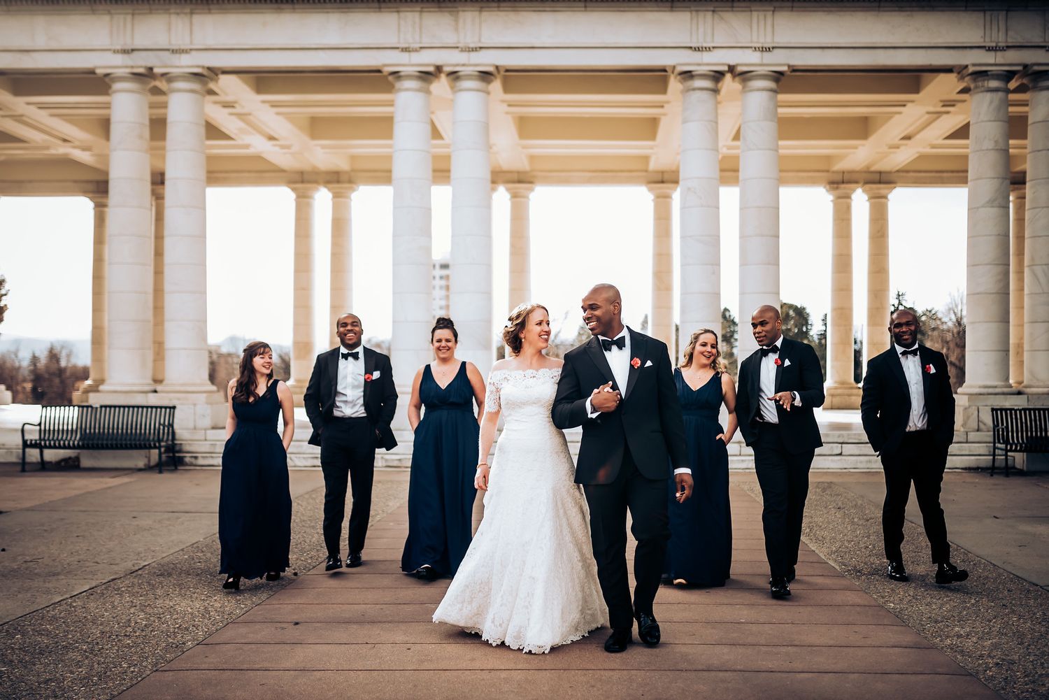 Wedding party in navy and white attire posing together under elegant stone columns at sunset.