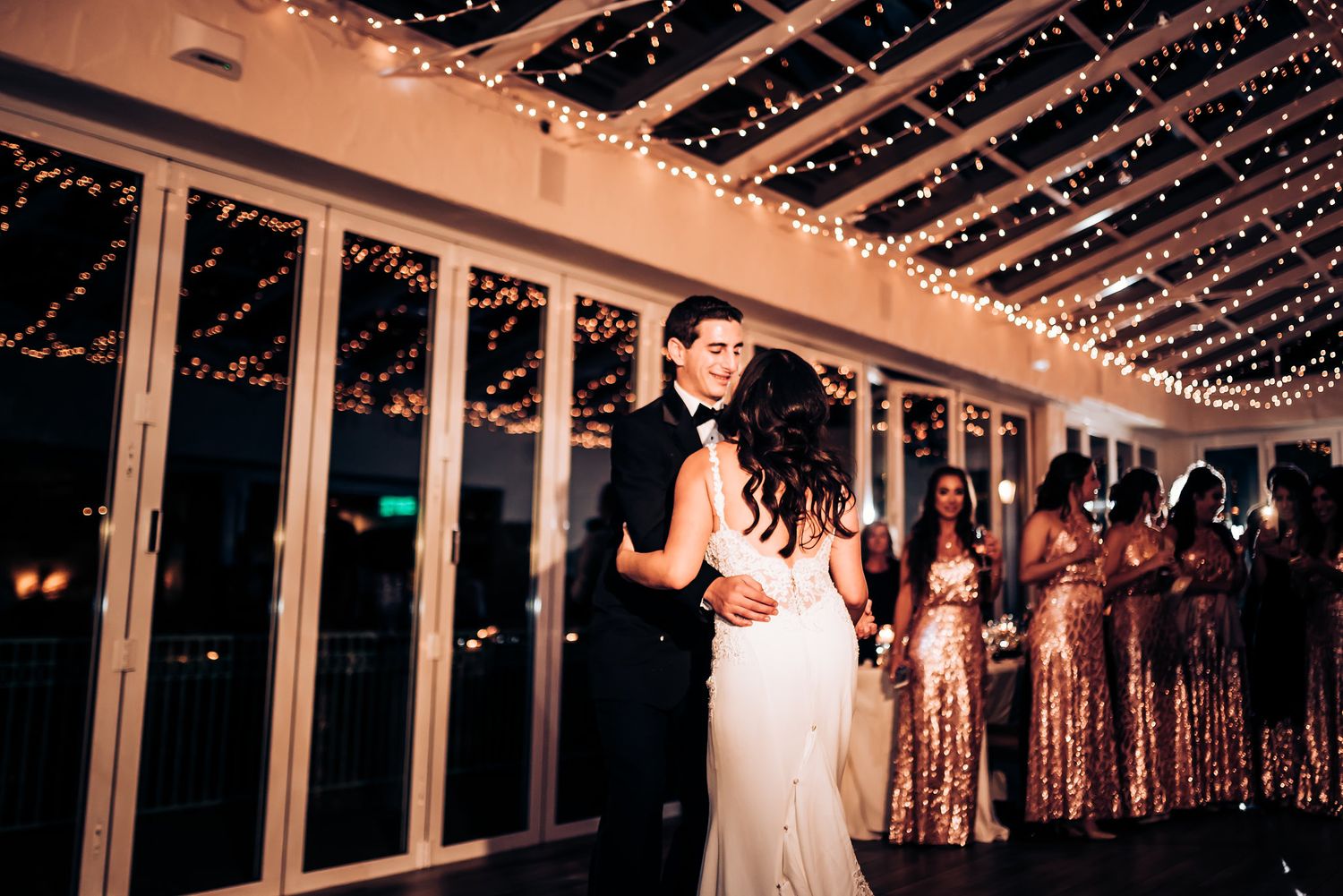 Couple shares intimate dance while bridesmaids in sequined dresses watch from background.