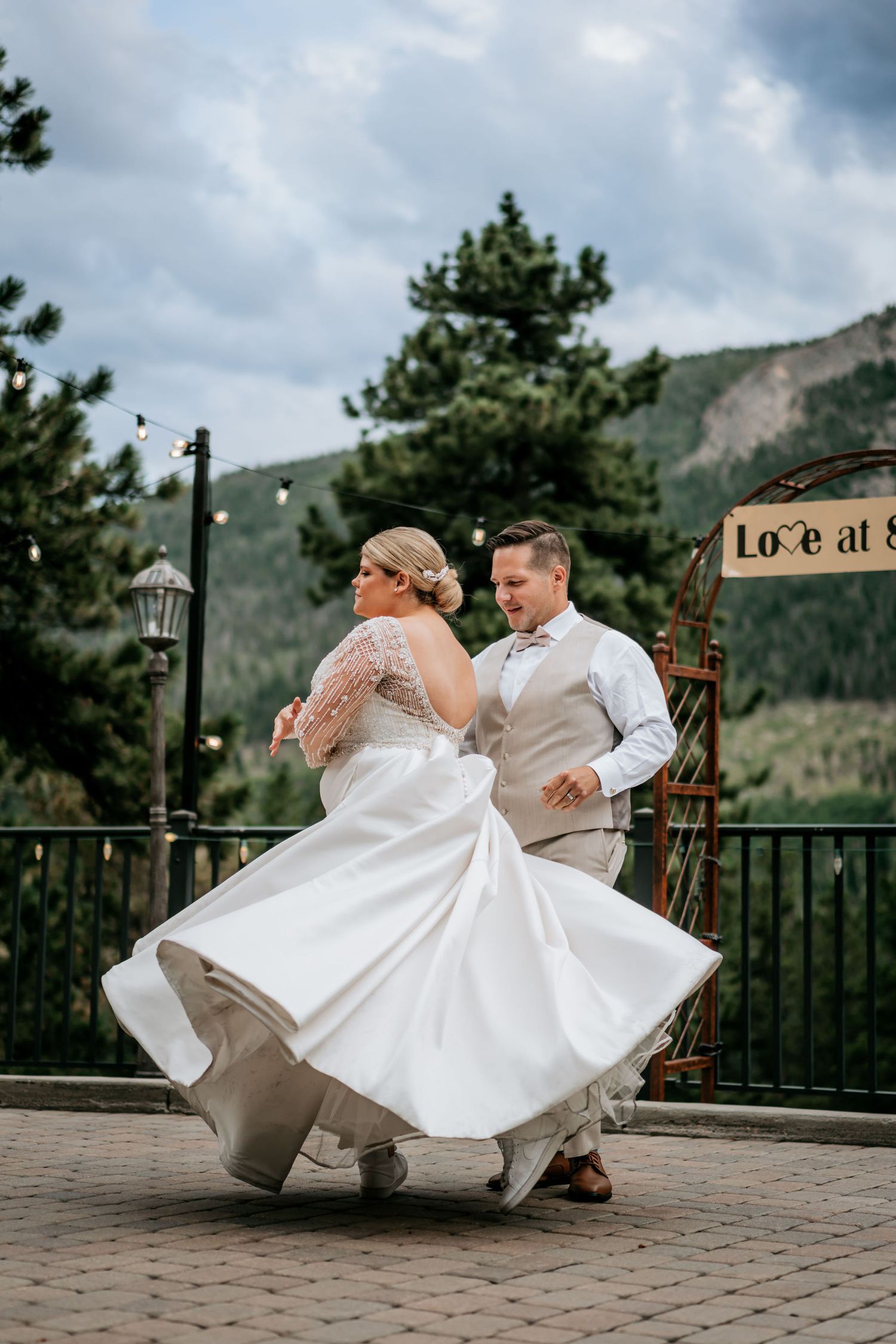 A wedding couple dances joyfully on a mountain deck with the bride's white dress twirling in motion.